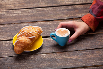 Wall Mural - Female hands holding cup of coffee with croissant on wooden tabl