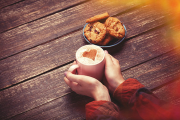 Wall Mural - Female hands holding cup of coffee and cookies on wooden table.