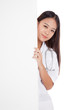 © halfbottle - Young Asian female doctor peeking from behind blank sign billboa