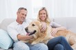 © WavebreakMediaMicro - Smiling couple petting their golden retriever on the couch