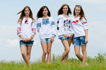 group of 4 beautiful young women girlfriends in folk embroidery dresses having fun happy smiling standing in green field & looking at camera on blue sky summer outdoors background portrait image