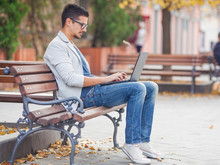 Young Man Sitting On The Park Bench With Laptop On His Lap