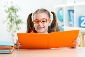 Wall Mural - Happy funny child girl in glasses reading a book in primary