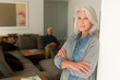 © Westend61 - Portrait of senior woman leaning at a wall while her husband sitting on the couch in the background