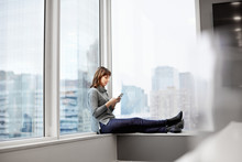 A Woman Holding A Smart Phone, Sitting On A Window Ledge.