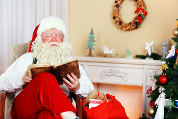 Santa Claus sitting with book in comfortable chair near fireplace at home