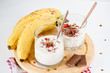 Two glasses with banana yogurt topped with granola, milk chocolate and goji berries on round wooden plank with bananas and milk chocolate bars on red napkin