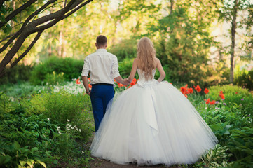 The bride and groom are in the park holding hands around the beautiful flower beds of poppies and white flowers