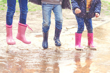 Low Section Of Three Children In Wellington Boots Jumping In A Puddle