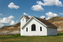 An Old White Church Nestled In A Ghost Town Among The Hills With A Bright Blue Sky And Puffy Clouds In The Summer Time