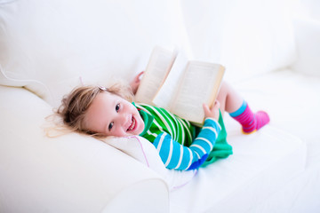 Wall Mural - Little girl reading a book on a white couch