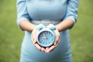 Asian pregnant woman holding an blue alarm clock, grass backgrou