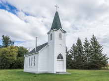 Horizontal Image Of A Beautiful Quaint Little Country Church Sitting On A Green Lawn Surrounded By Spruce Trees Under A White Puffy Cloud Filled Sky In The Summer Time.