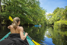 Woman Kayaker Paddling Down A Beautiful Jungle River With Two People In A Canoe On A Gorgeous Day. Lots Of Copy Space And View From Behind