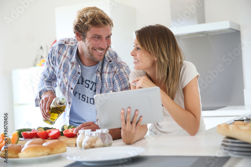 Obraz na plátně Cheerful couple in kitchen cooking dinner, using tablet