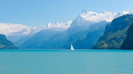 Canvas Print - Magnificent panorama of mountains with snowy peaks. On  surface of lake sailboat sails. Cloudless blue sky and a few clouds in the mountains