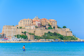 Wall Mural - Man relaxing in sea water and view of citadel with houses in Calvi port in background, Corsica island, France.