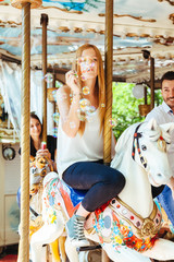 On a summer day a group of friends having fun in the shade on a carousel with horses and blowing soap bubbles. In the foreground a young woman in the air while blowing many colorful soap bubbles