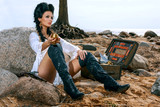 Beautiful pirate woman sitting near treasure chest on the beach with a golden goblet in her hand