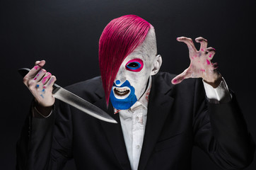 Wall Mural - Clown and Halloween theme: Scary clown with pink hair in a black suit with a knife in his hand on a dark background in the studio