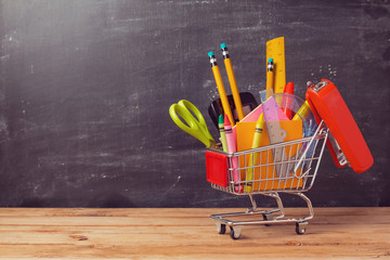 Shopping cart with school supplies over chalkboard background. Back to school sale concept