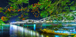 Trang Tien Bridge lit up at night reflected underwater beside sparkling colors are flamboyant flowering stems in summer weather, beneath the boat leaving dock carrying travelers.