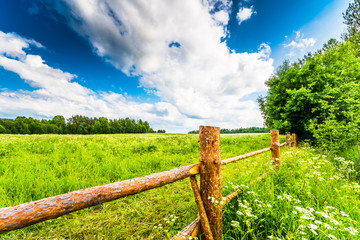 Field in the forest enclosed by a wooden fence on a background of the cloudy sky