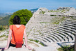 Tourist girl with an orange t-shirt sitting on one of the steps of the Segesta theather, West Sicily, and looking at the view