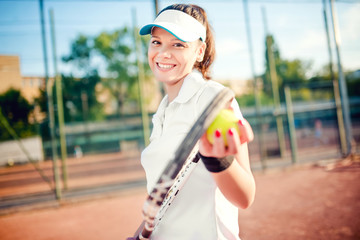 Portrait of woman playing tennis, holding racket and ball. Attractive brunette girl wearing white t-shirt and cap on tennis court