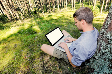 man is sitting against a tree in the forest, working with his la