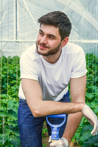 Young sitting between rows of plants, working in a greenhouse. Poster