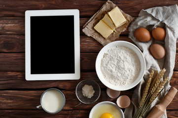 Poster - White tablet computer with blank screen with ingredients for making pancakes or cake - flour, egg, butter, milk on the wooden background. top view. rustic style. free space for text . Copy space
