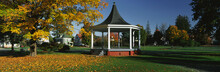 This Is The Town Square. In The Center Is A Gazebo. It Is Located On Main Street. There Is Autumn Foliage In A Park Setting. We See Victorian Style Houses In The Background. The Gazebo Has White Columns And Steps Leading Up To Its Landing. It Has A Black Roof Covering It.