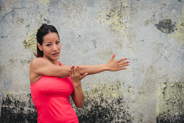 Wall Mural - Fitness female athlete stretching arm and shoulder. Sporty woman exercising and working out against a wall outside.