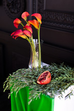 Bouquet Of Red Calla Lilies In A Glass Vase With A Pomegranate And Eucalyptus Stands On The Bedside Table On A Black Background 