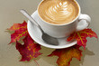 Autumn Coffee Latte with Orange and Red Fall Leaves. A hot latte with a design in the foam sits on a white plate with a spoon on a glass table with red, orange, and yellow autumn fallen leaves at cafe