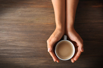 Sticker - Female hands holding a cup of coffee with foam over wooden table, top view