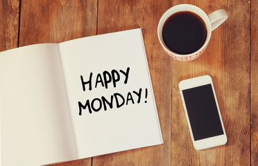 notebook with the phrase happy monday written on it, coffee cup and smart phone. filtered image.
