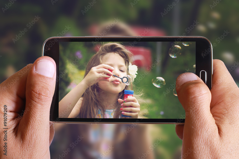 Male hand taking photo of Vintage Portrait of beautiful girl blowing bubbles with cell, mobile phone. Kids, childhood, family concept. - obrazy, fototapety, plakaty 