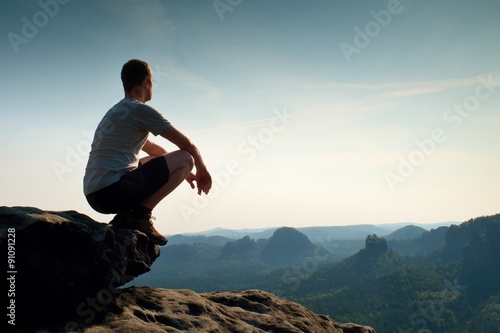 Cuadros en Lienzo Young man in black sports pants and grey shirt is sitting on cliff's edge and l