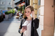 Young beautiful business woman standing on the street with coffee and smart phone. Smiling and talking on the phone.
