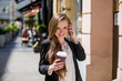 Young beautiful business woman standing on the street with coffee and smart phone. Smiling and talking on the phone.
