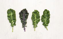 Three Leaves Of Fresh Green Kale And One Of Red Kale On White Table