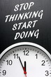 The phrase Stop Thinking Start Doing in white text on a blackboard above a clock with the hands pointing towards twelve or midnight