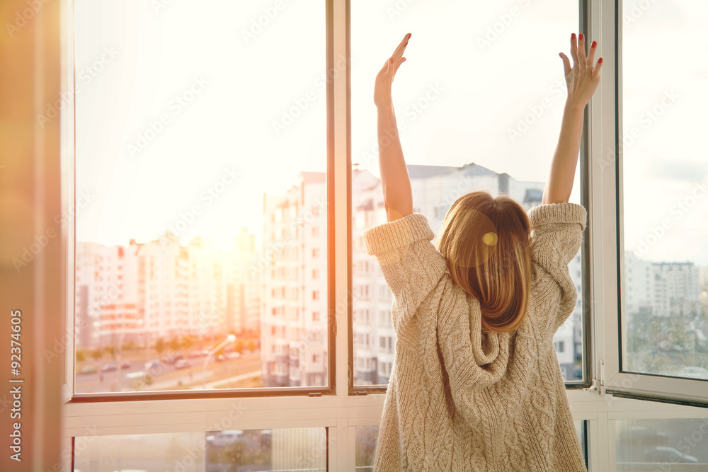Woman near window raising hands facing the sunrise at morning - obrazy, fototapety, plakaty 