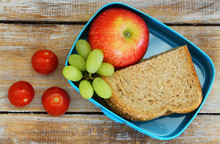 Lunch Box Containing Brown Bread Sandwich, Red Apple, Grapes And Cherry Tomatoes On Rustic Wooden Surface
