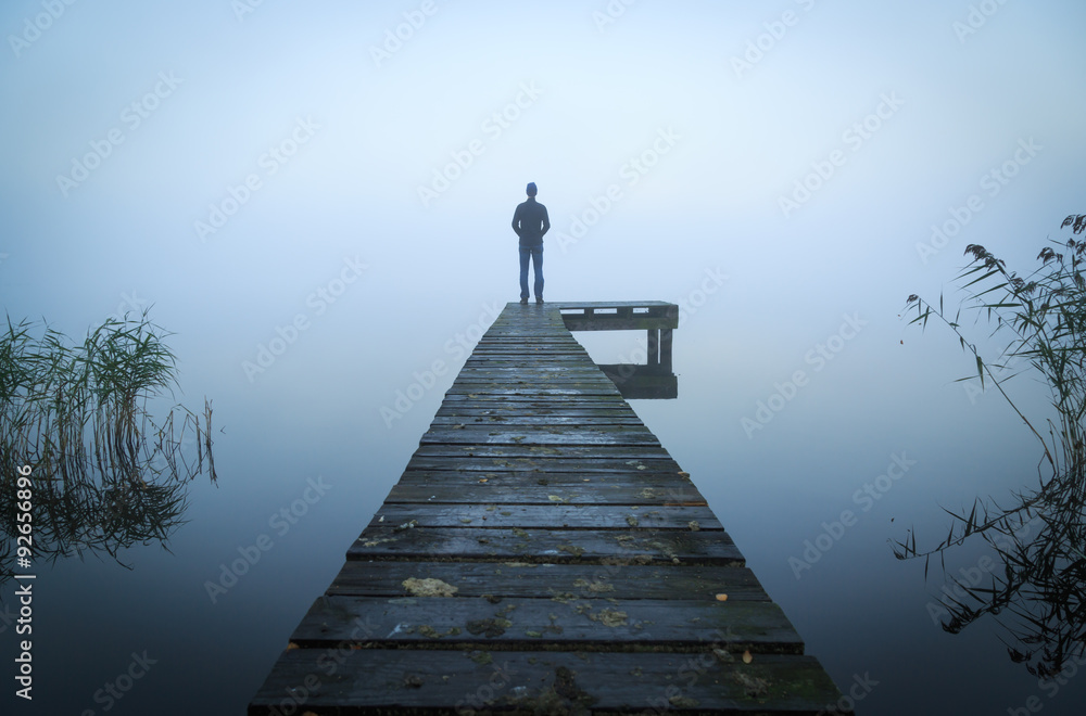 Man standing on a jetty at a lake during a foggy, gray morning. - obrazy, fototapety, plakaty 