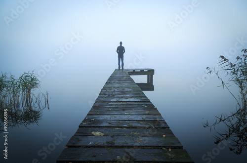 Fotografía Man standing on a jetty at a lake during a foggy, gray morning.