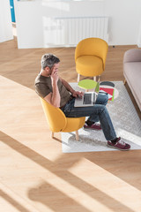 Top view  of a middle aged grey hair man with beard in a stylish vintage living room with wooden floor. He is sitting in a design orange chair with his laptop on his knees He is wearing casual clothes