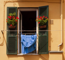 Italian Colors: Window With Open Shutters, Red Geranium Vases And Bue Laundry Hanging In The Sun